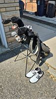 Full view of the golf stand bag loaded with multiple golf clubs including irons and woods, some with black furry headcovers, along with white golf shoes size 11.