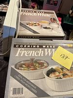Photo of Corning Ware French White cookware set box featuring white ceramic dishes with ribbed sides and glass lid, showcasing product use with food.