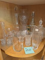 Crystal glassware collection displayed on wooden shelf, including several decanters, vases, and a large pitcher with cut glass patterns.