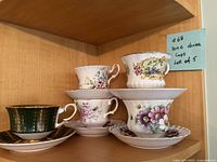 Five bone china cups with different floral and decorative designs stacked paired with matching saucers inside a wooden cabinet shelf. A sticky note labels them as '#69 Bone china Cups Lot of 5'.
