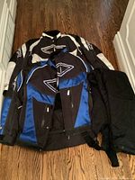 Two snowmobile jackets, one black and white and one black and blue, with black snowmobile pants laid out on a wooden floor