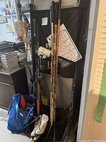 Full view of six hockey sticks, white ice skates, and coaching board displayed upright against a wall with a blue bag near the base.