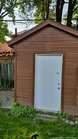 Photo of the exterior front wall of a wooden garden shed with a white door, surrounded by grass and trees in a backyard.