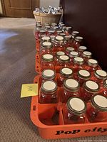 Multiple clear glass canning jars with silver metal screw-on lids arranged in two orange crates and one white tray on carpet. A wooden basket filled with additional metal lids is visible behind them.