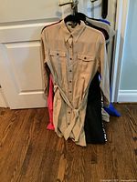 Photo showing a beige shirt dress with buttons, chest pockets, collar, and waist belt hanging on a rack among other dresses in various colors.