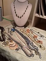 Overall view of thirteen assorted necklaces displayed on table and bust, showing variety in style, materials and length.