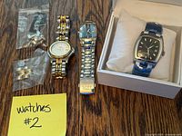 Photo showing three men's watches on wooden surface along with small bags containing extra watch band links. Two watches are used with visible band wear; one watch is boxed and new.