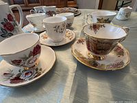 Photo of multiple fine bone china teacups and saucers with floral designs and gold trim on a table.