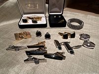 Overview of all men’s jewelry items including multiple cufflinks, tie clips, and a rhinestone stretch bracelet arranged on a beige fabric.