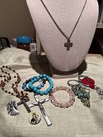 Photo showing assortment of religious jewelry including rosary, bead bracelets, cross pendants, charms, and a keychain on a surface.