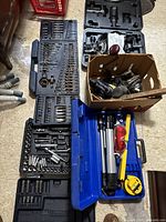 Photo showing multiple tool cases opened to reveal socket wrenches, drill bits, power tools in a box, and accessories such as goggles and measuring tape.
