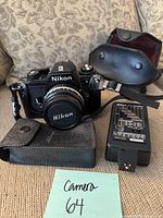 Front view of Nikon EM camera with attached Nikon Series E 50mm lens, lens cap on, black leather case in front, and Speedlight SB-E flash beside it with leather protective cover behind.