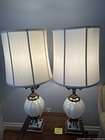 Two white glass and brass metal table lamps with white fabric shades placed on a wooden table, showing overall condition and style.
