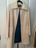 Front view of the off-white long coat hanging, showing button closure and visible texture of fabric. Blue lining is visible inside.