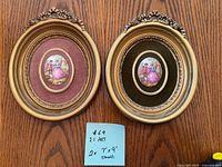 Two oval framed art pieces on a wooden floor showing detailed gold-toned frames and contrasting maroon and dark green mats, each having romantic Victorian couple images.