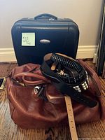 Photo showing the black hard-shell suitcase behind a brown leather duffel bag with three black belts draped over the duffel bag, showcasing the overall lot items.