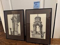 Set of two framed architectural artwork prints with black and white detailed designs, standing side by side on floor against a wall.
