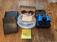 Photo showing two black leather wallets, Guess sunglasses in a case, Roots eyeglasses in a white case, and blue binoculars with a pouch all arranged on a wooden floor.