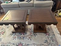 Image showing two brown wooden side tables with square tops and decorative bases placed on a floral carpet in front of a sofa.