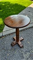 Side angle view of the wooden round side table showing the top, pedestal column, and four-foot base on asphalt outside.