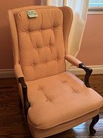 Full view of the traditional wing chair showing peach tufted upholstery, wooden arms with dark finish, and carved legs on hardwood floor.