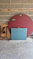 Photo of two portable folding tabletops, one round maroon and one rectangular blue-grey, with a set of metal folding table legs beside them.