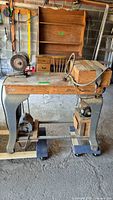 Front view of vintage powered table saw showing wooden tabletop, grindstone attachment on left side, electric motor mounted below, and foot pedals.