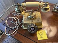 Front and side view of vintage rotary phone on wooden table, showing brass base with floral detailing and ivory handset with cord coiled beside it.