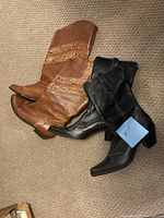 Two pairs of women's boots on carpet: brown taller boot with decorative stitched bands and black ankle boot with decorative stitching on toe and heel.