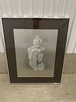 Framed grayscale artwork showing a seated nude woman with her chin resting on her knees, viewed from the front.