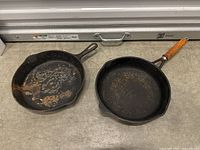 Two cast iron skillets side by side showing worn cooking surfaces; left skillet heavily worn, right skillet with wooden handle.