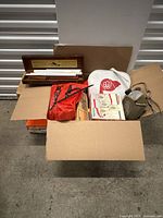 Full top-down view of an open cardboard box containing the various items including the wooden cribbage board, white Olympics bag, glassware box, and canvas canteen.
