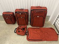 Five pieces of red luggage including three upright suitcases, duffle bag, and garment bag all displayed on a concrete floor.