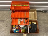 View of open tackle box showing orange tray with compartments filled with various fishing accessories including small ropes, a bobber, and hooks. The bottom compartment contains fishing line, fishing bobbers, wire cutters, packaged hooks, and other small fishing-related tools.