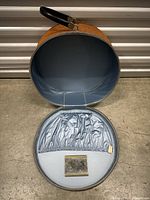 Open round carrying case showing light blue interior lining with elastic pocket and small metal plaque/mirror