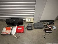 Photo of vintage electronics lot items arranged on the floor including cassette players, clock radio, tape accessories and cables