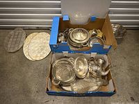 Two cardboard boxes filled with assorted vintage silver-plated and glass kitchen serving items including trays, a teapot, and decanter.