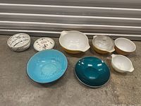 Assorted vintage Pyrex and ceramic bowls in turquoise, yellow, and white, arranged on floor.
