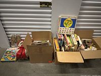Multiple cardboard boxes filled with assorted vintage toys and board games alongside loose toys on the floor against a closed door.