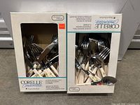 Two boxed sets of Corelle Coordinates flatware, showing packaging and contents inside boxes: forks, knives, spoons with floral patterned cream handles.