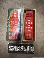 Red handheld Merlin game with twelve white circular buttons and four labeled rectangular buttons, plus original colorful box.