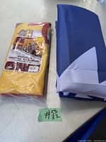 Two folded flags placed on a table, one in original packaging showing Spain flag colors and label, the other folded in blue and white fabric.
