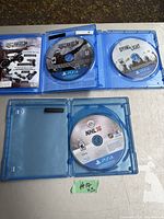 Top down view of the three PS4 game discs inside their blue cases showing titles Homefront: The Revolution, Dying Light, NHL 16. Homefront case includes manual.