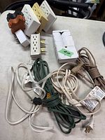 Photo showing four extension cords in different colors and seven white wall taps arranged on a surface.