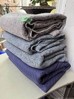 Stacked view of four folded moving blankets: three grey felt-type blankets and one navy-blue quilted blanket at bottom.