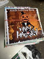 Stack of adult coloring books bound with two rubber bands, showing the front cover of a Halloween-themed book titled 'Adult Coloring Book Happy Halloween' having an illustrated pumpkin-headed character in a tuxedo.