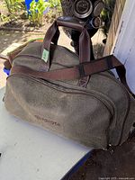 Photo of olive green Toyota duffle bag with brown handles and shoulder strap, showing front zippered pocket and Toyota logo.