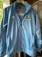 Front view of a light blue Adidas jacket with zipper, showing Adidas logo on right chest and an embroidered emblem on left chest.