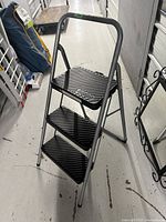 Photo shows a three-step folding step ladder with silver metal frame and black ribbed plastic steps, standing on a floor indoors.