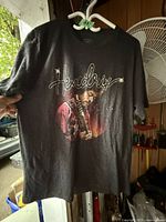 Front view of black Hendrix T-shirt held by hand showing colorful graphic of Jimi Hendrix and the text "Hendrix".